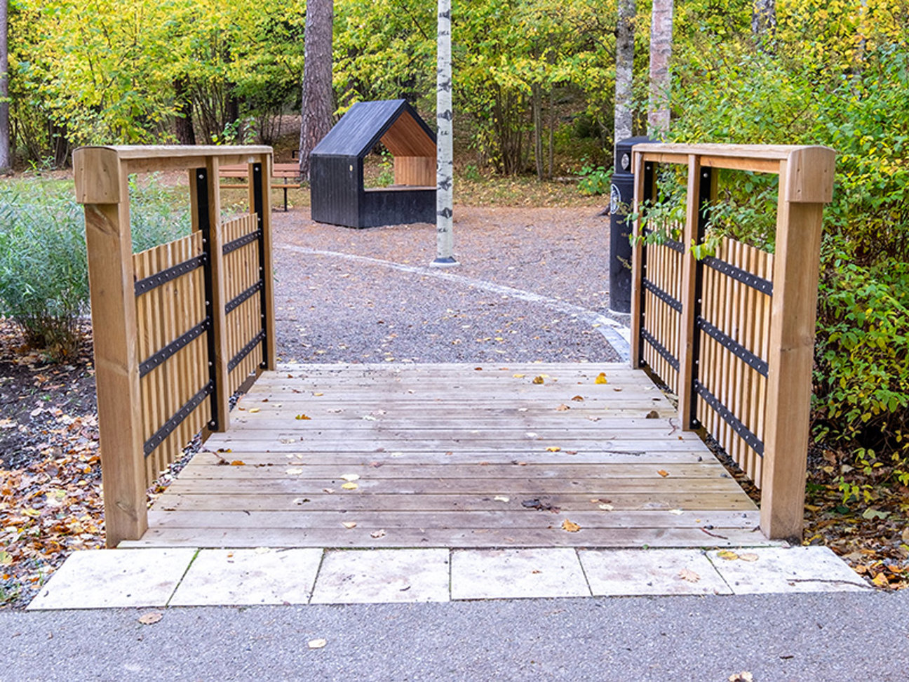 Träbro med staket, grusväg och liten byggnad omgiven av träd.