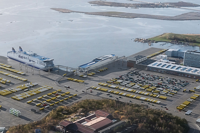 Flygfoto av hamnområde med fartyg, parkeringsplatser och byggnader vid vatten.