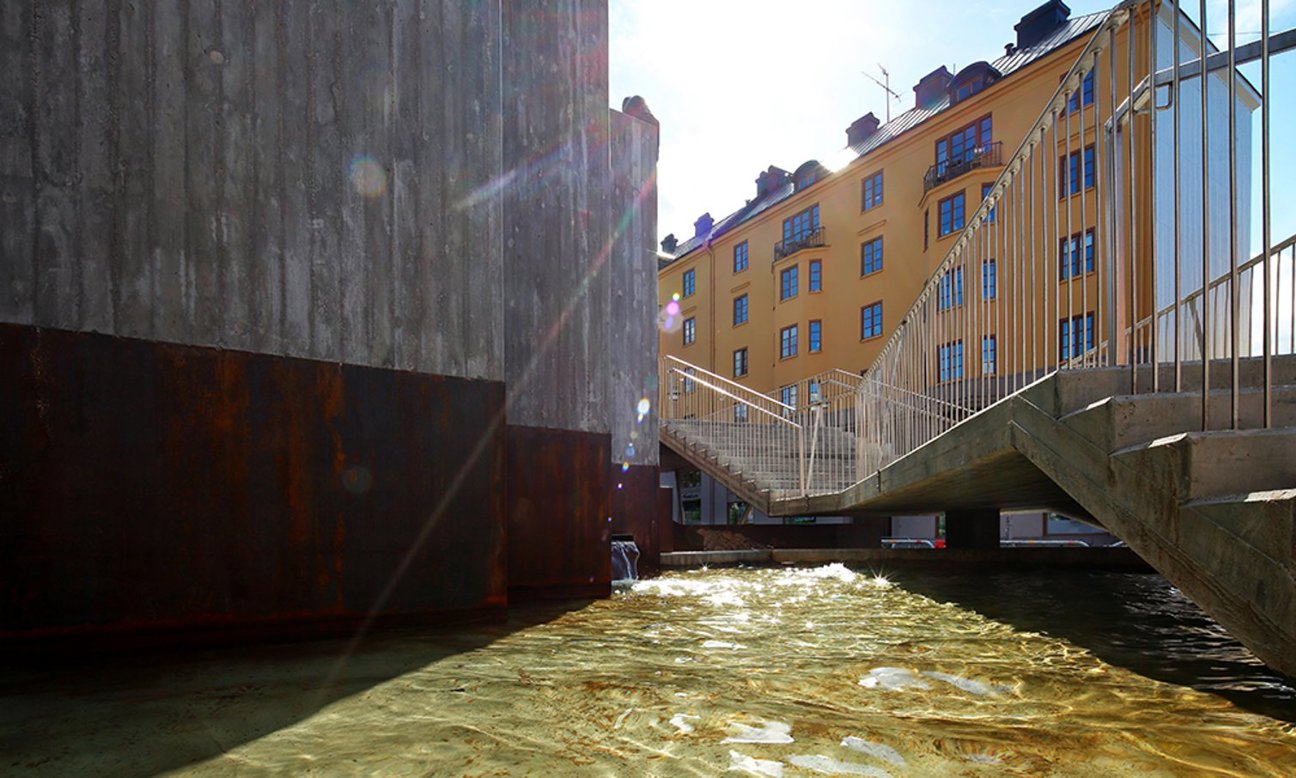 En bro över vatten med gula byggnader och betongstruktur i bakgrunden.