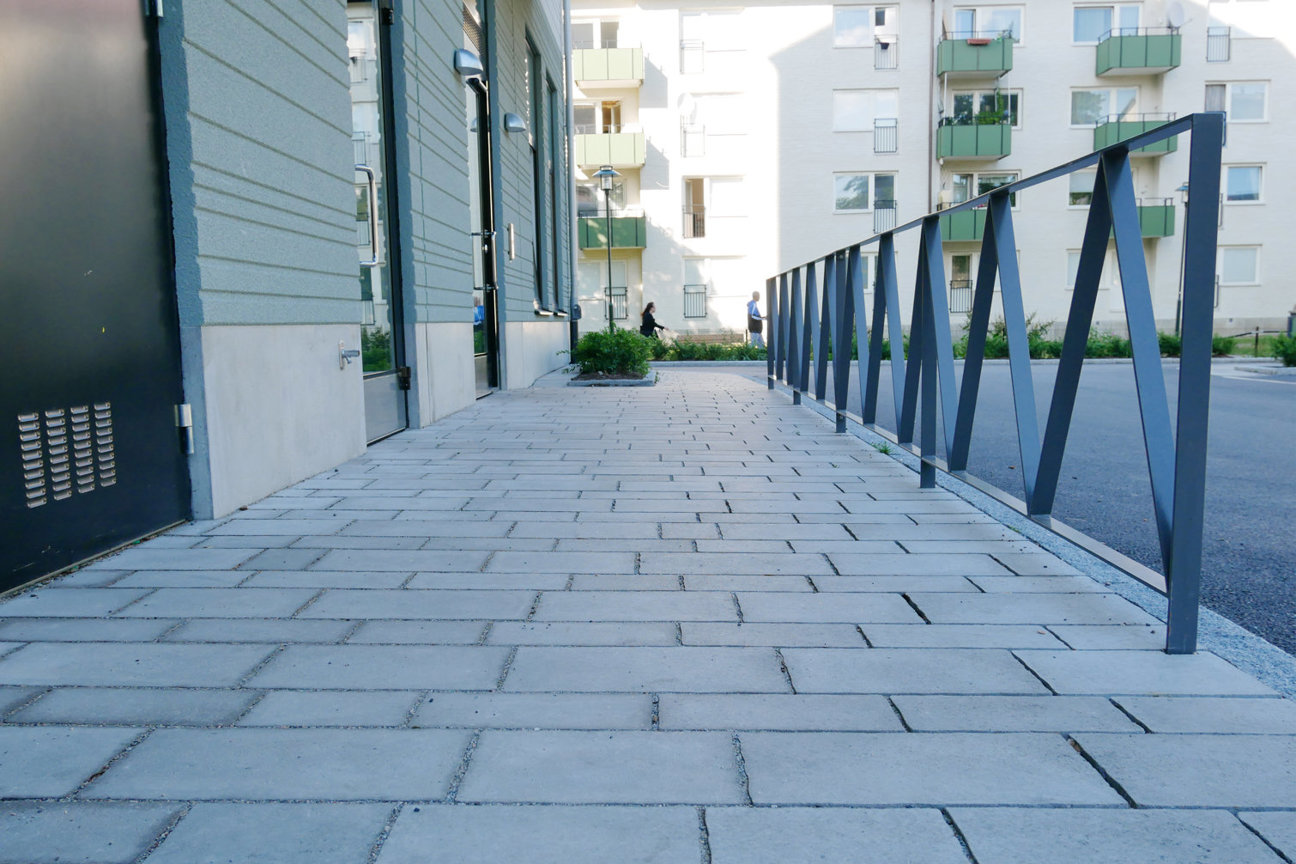 Stenlagd gång mellan byggnader med metallräcke och flerfamiljshus i bakgrunden.