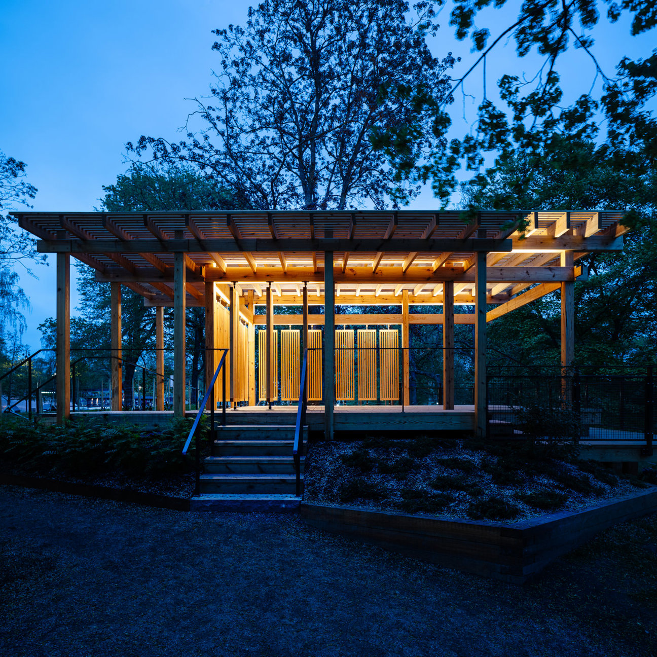 En träpaviljong med trappa och belysning omgiven av träd vid skymning