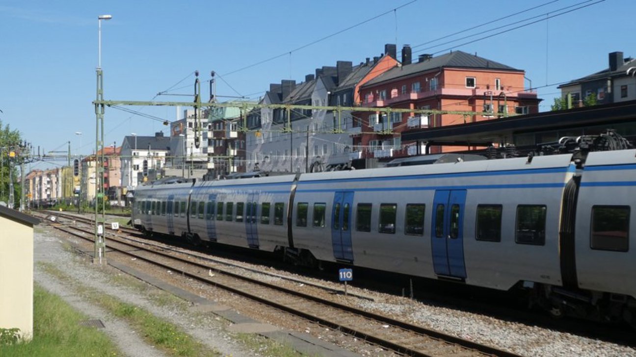 Tåg på järnvägsspår med flerfamiljshus i bakgrunden under klar himmel.