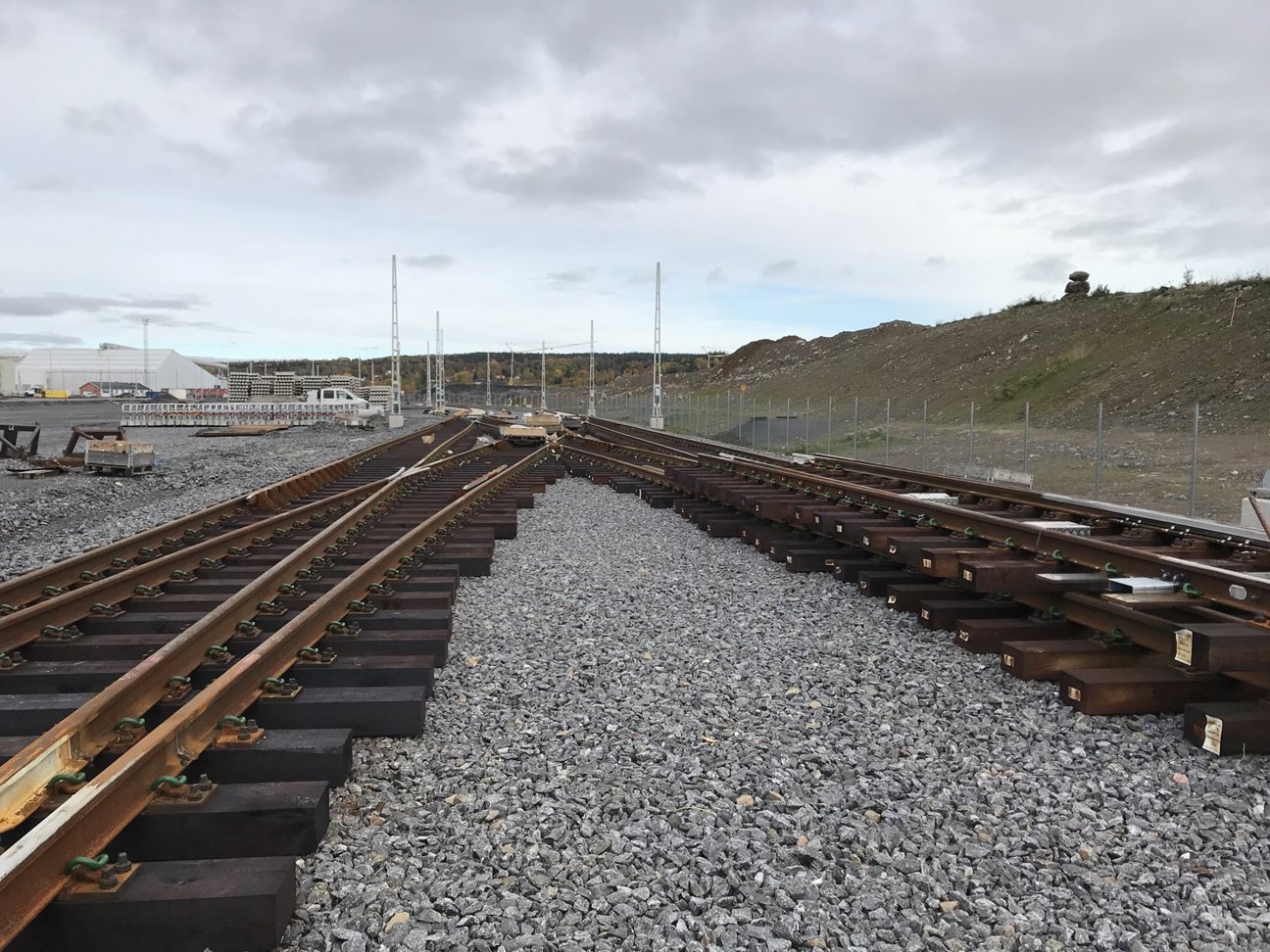 Järnvägsspår med grus och industriell anläggning under molnig himmel.