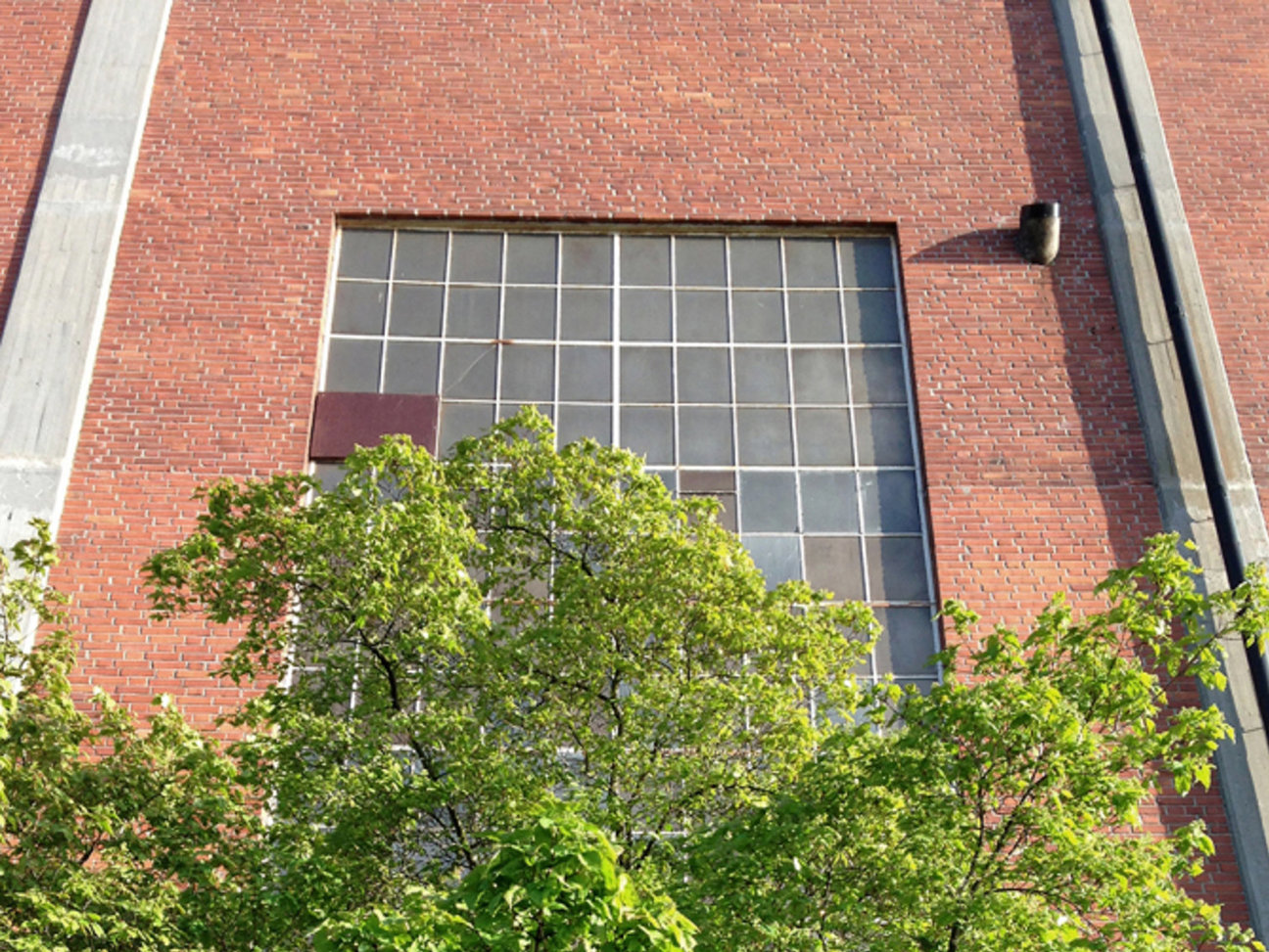 Röd tegelvägg med stort rutigt fönster och gröna träd i förgrunden.
