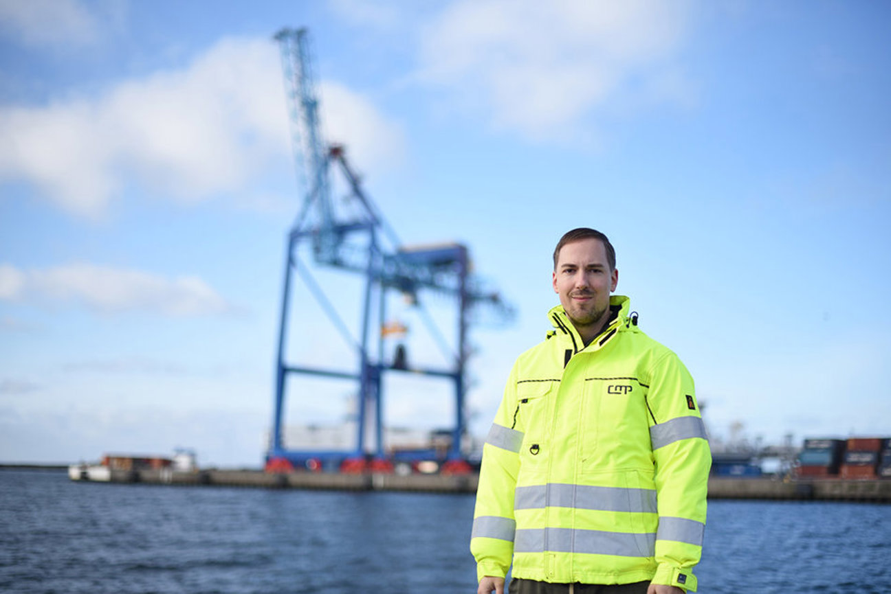 En person i neongul säkerhetsjacka framför hamnkranar vid vatten under blå himmel.