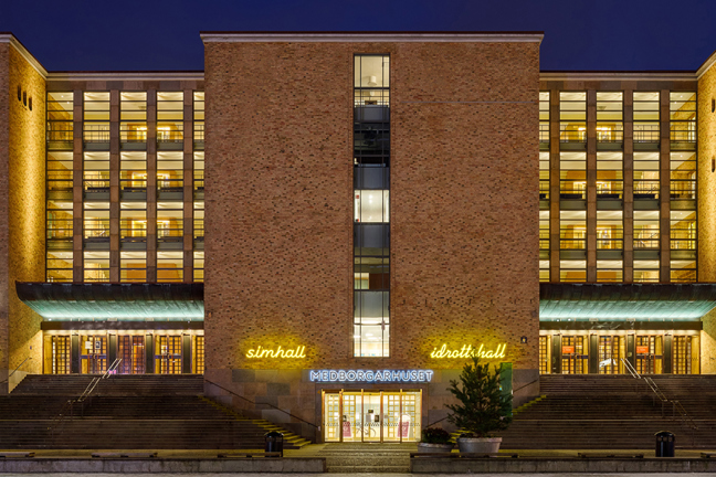 Nattbild av en tegelbyggnad med belysta fönster och neonreklamer för stadsbibliotek och Kulturskolan