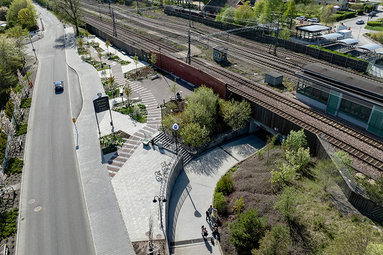 Flygvy över gata, järnvägsspår, byggnader och några personer på gångväg.