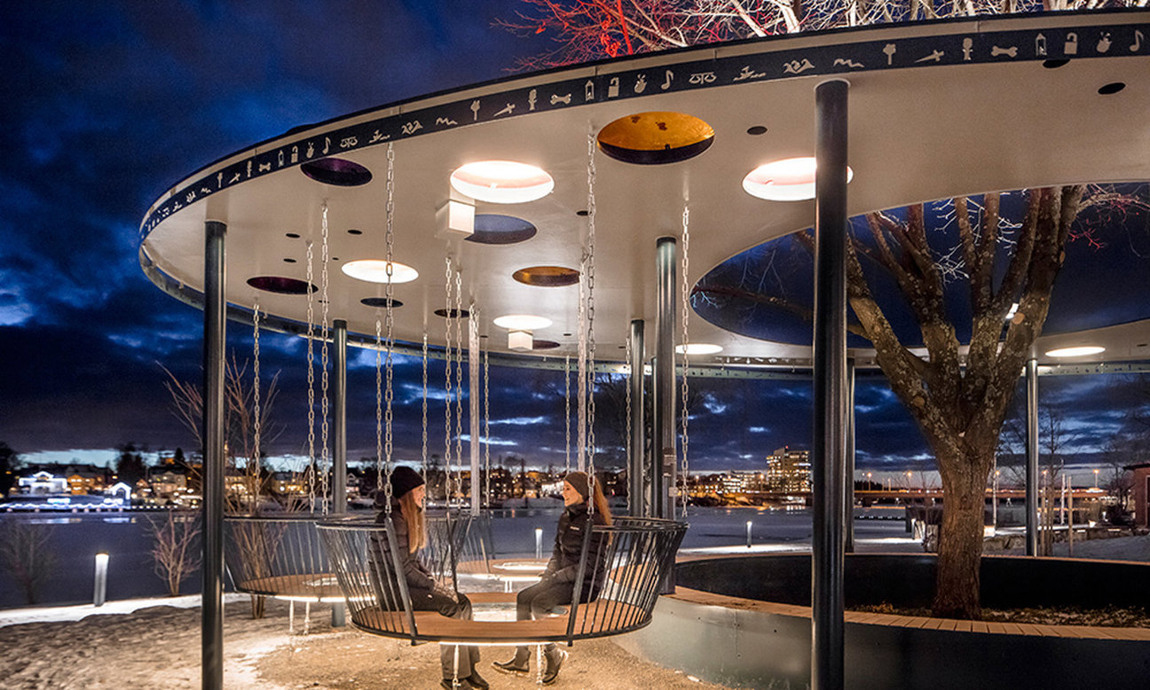 Cirkulär byggnad med hängande svängar, två personer på svängarna, träd och vatten i bakgrunden vid natt.