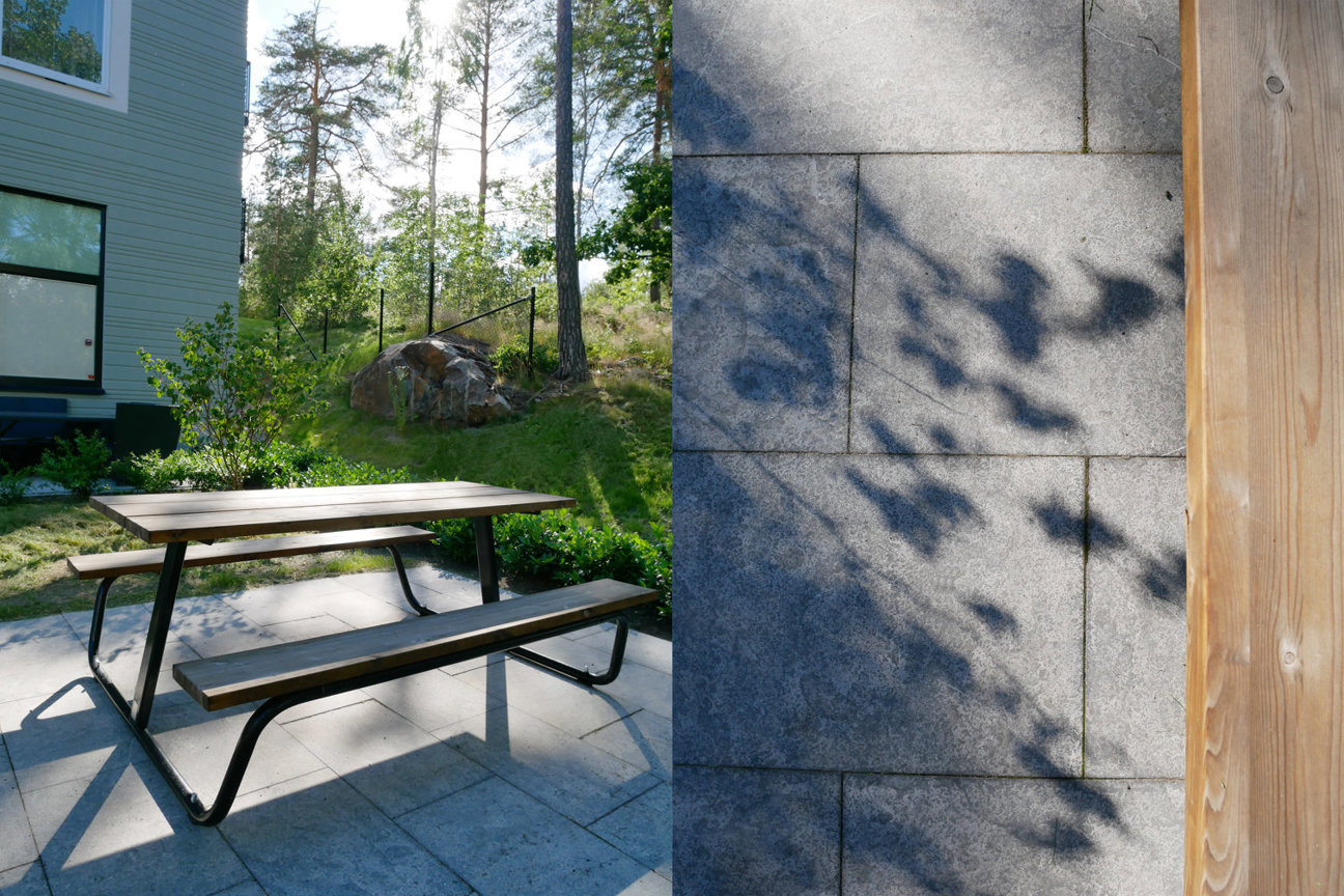 Utomhusplats med picknickbord, hus, träd och stenmur med skuggor.