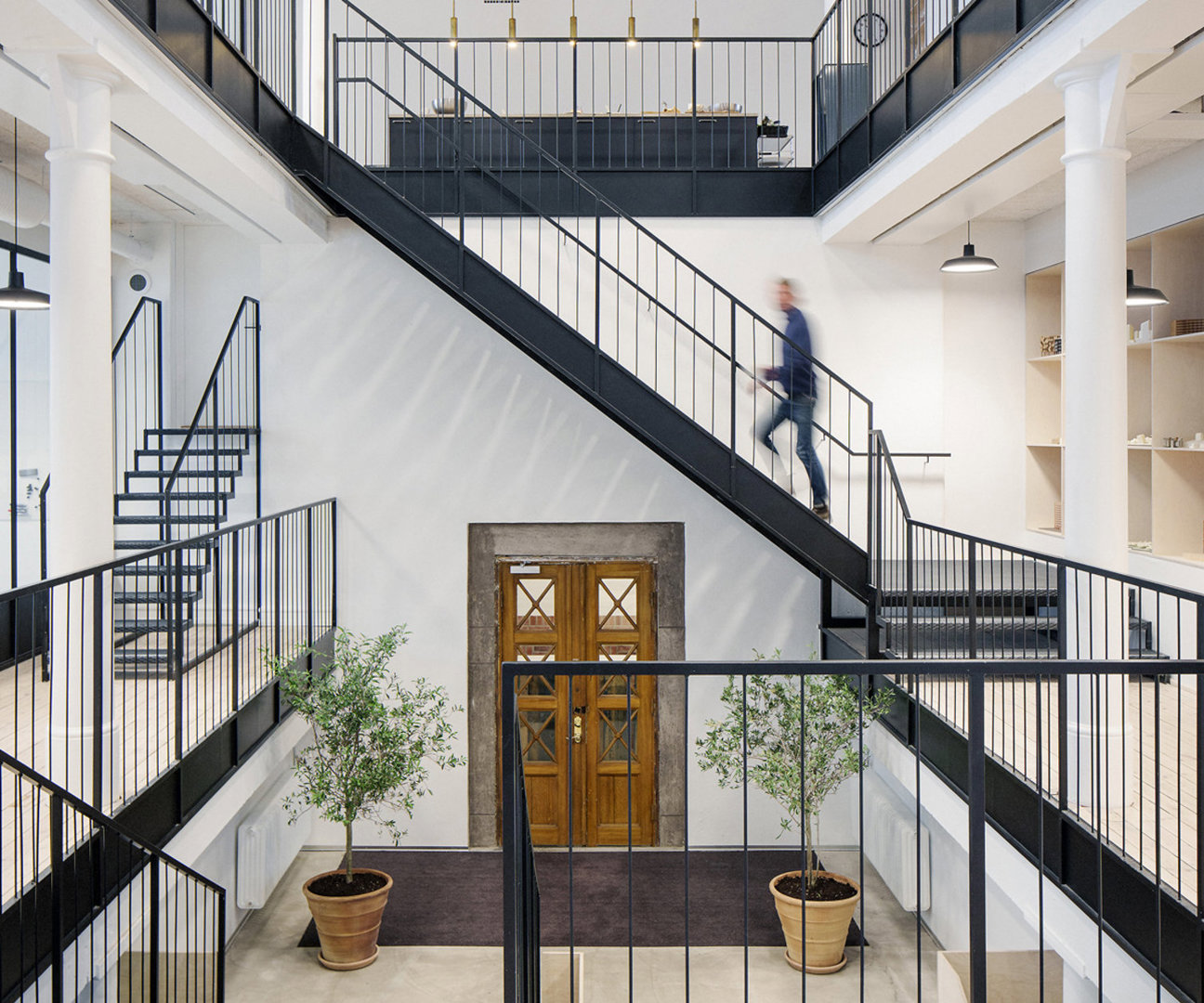 Interiör med svarta trappor, vita väggar, trädörrar och krukväxter. En person på trappan.
