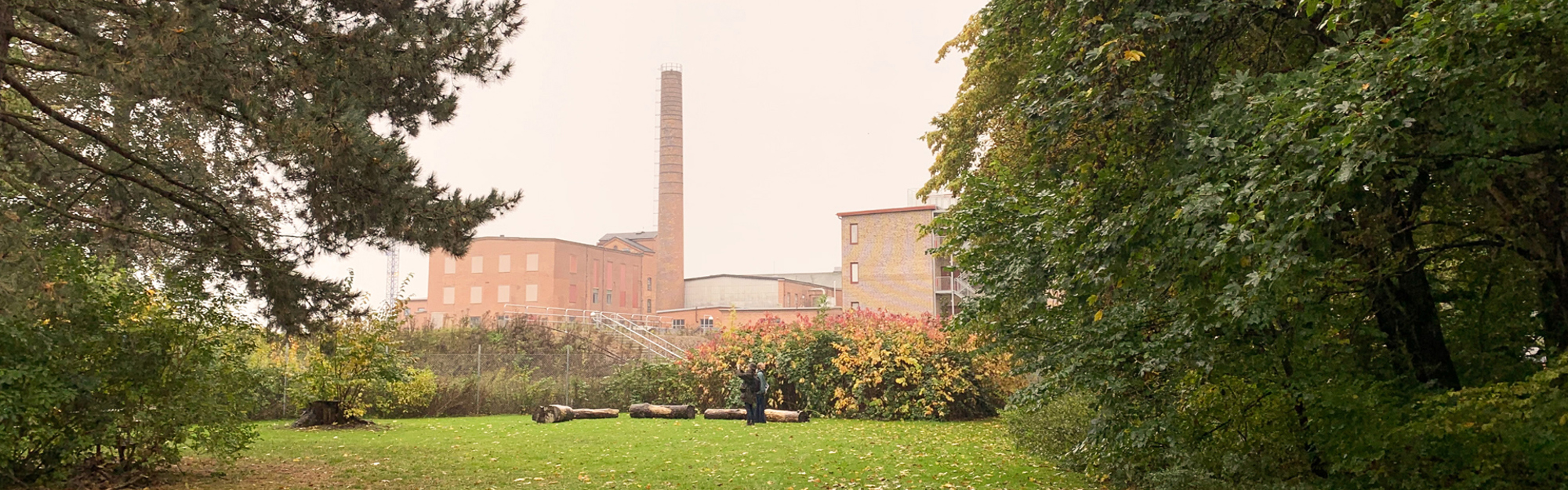 En byggnad med hög skorsten omgiven av träd och gräs, två personer står vid timmerstockar.