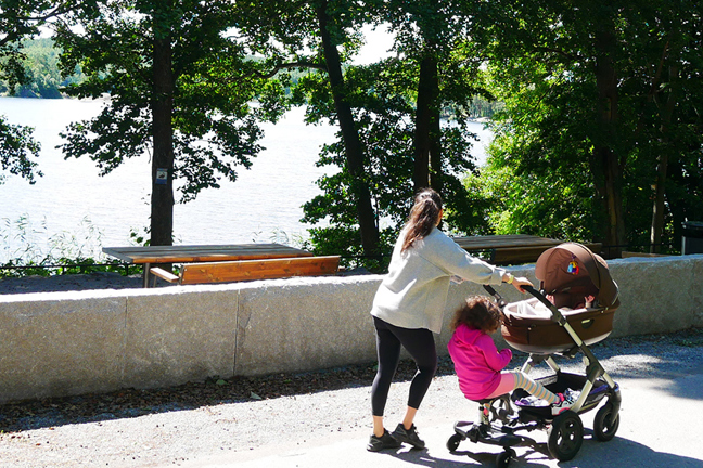 En kvinna och ett barn vid en barnvagn på en promenadväg vid en sjö med bänkar och träd.