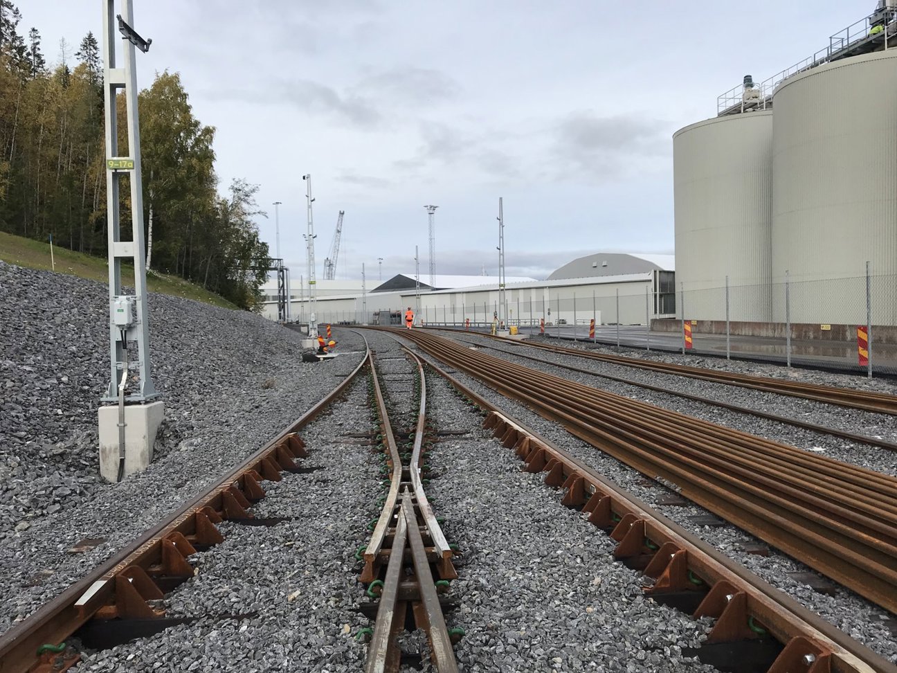 Järnvägsspår med grus, stora tankar och en person i orange väst i bakgrunden.