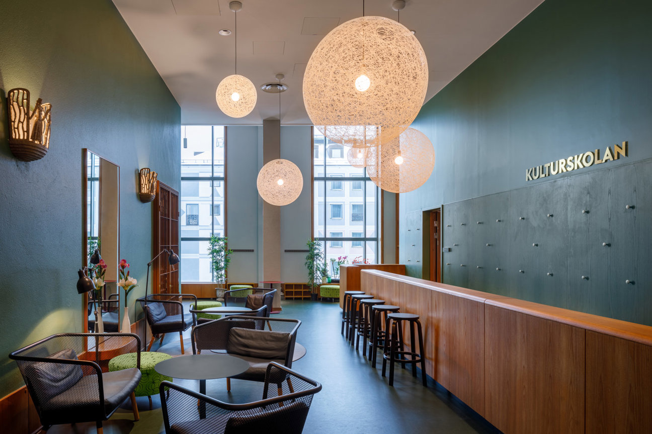 En modern interiör med gröna väggar, stora fönster, sfäriska lampor, stolar och bord. Texten 'KULTURSKOLAN' syns på en vägg.
