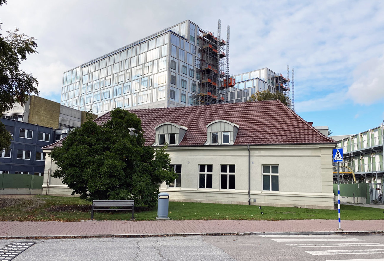 Ljust byggnad med röd tegeltak, moderna höghus under byggnad i bakgrunden, bänk och gångmarkering i förgrunden.