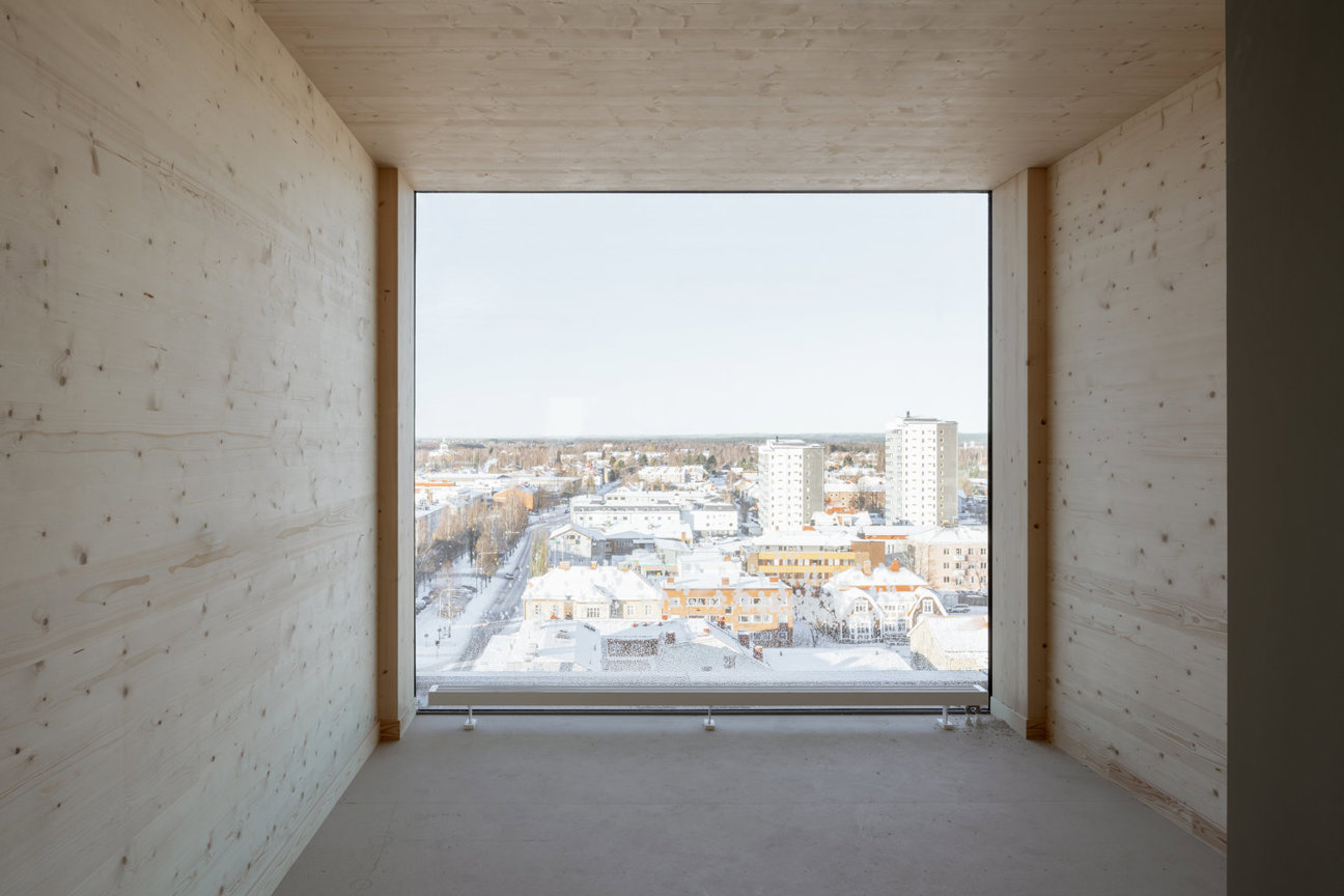 Ett rum med ljusa träväggar och stort fönster som ger utsikt över en snöig stadslandskap med flera byggnader.
