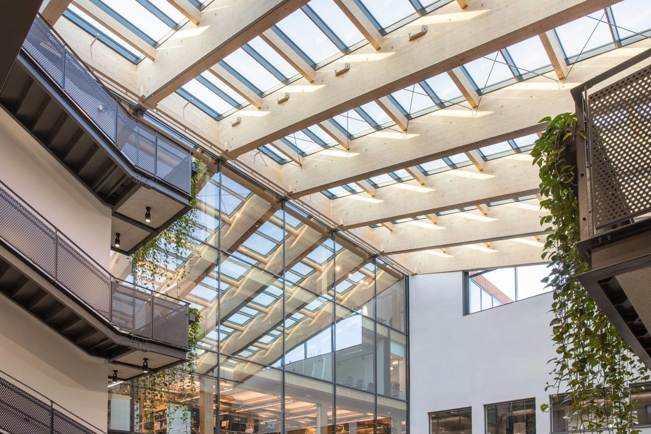 Inre av en modern byggnad med glastak, träbalkar, glasväggar och gröna växter.