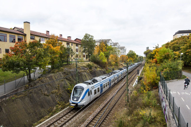 Tåg på järnvägsspår omgivet av hus och träd med höstfärgade löv. En person cyklar på en väg bredvid.