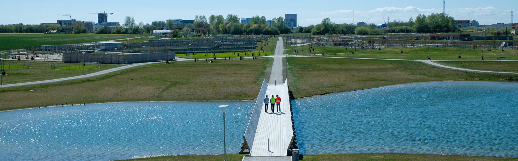 Tre personer på en bro över en sjö med gräs och byggnader i bakgrunden.