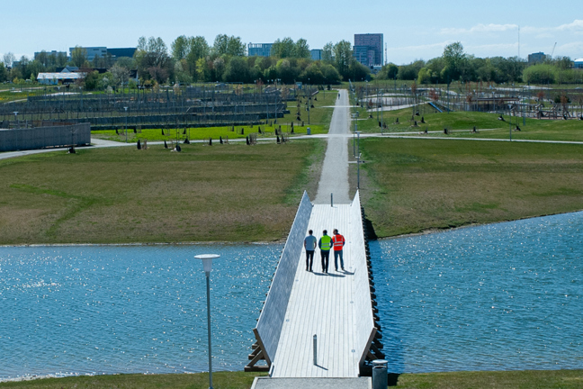 Tre personer på en bro över en sjö med gräs och byggnader i bakgrunden.