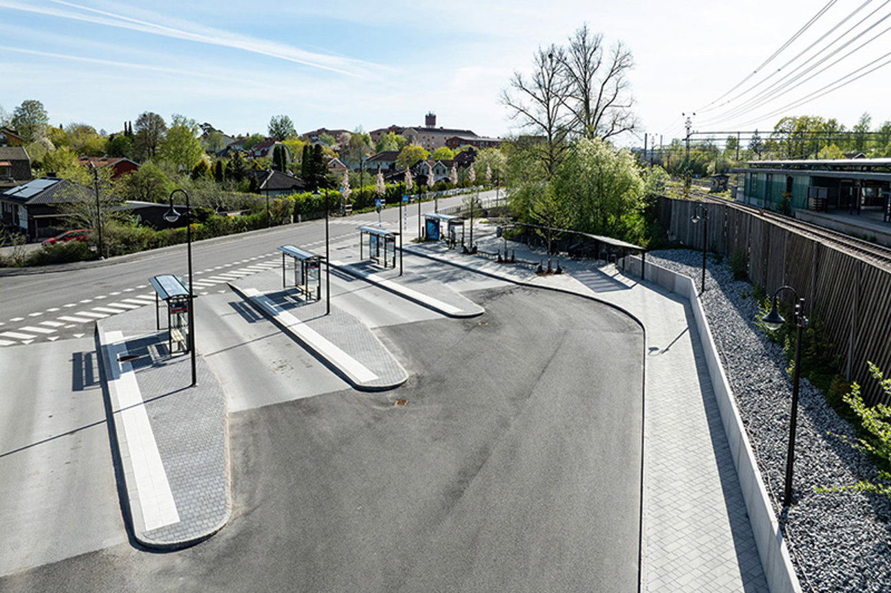 En vägkorsning med busshållplatser, träd och bostäder i bakgrunden.