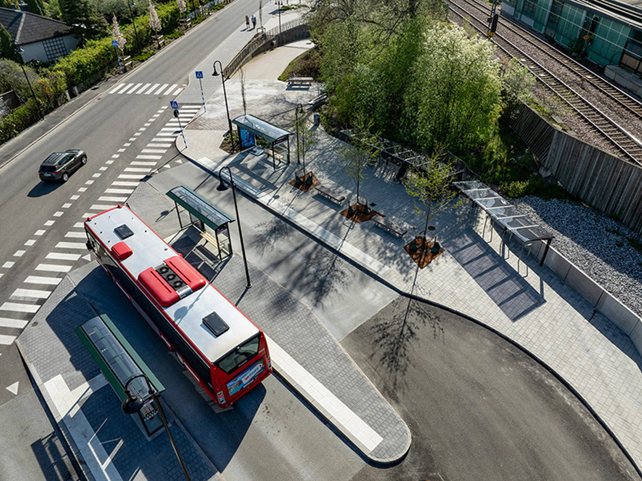 Luftbild på busshållplats med röd buss, väg, övergångsställe, träd, bänkar och järnvägsspår.