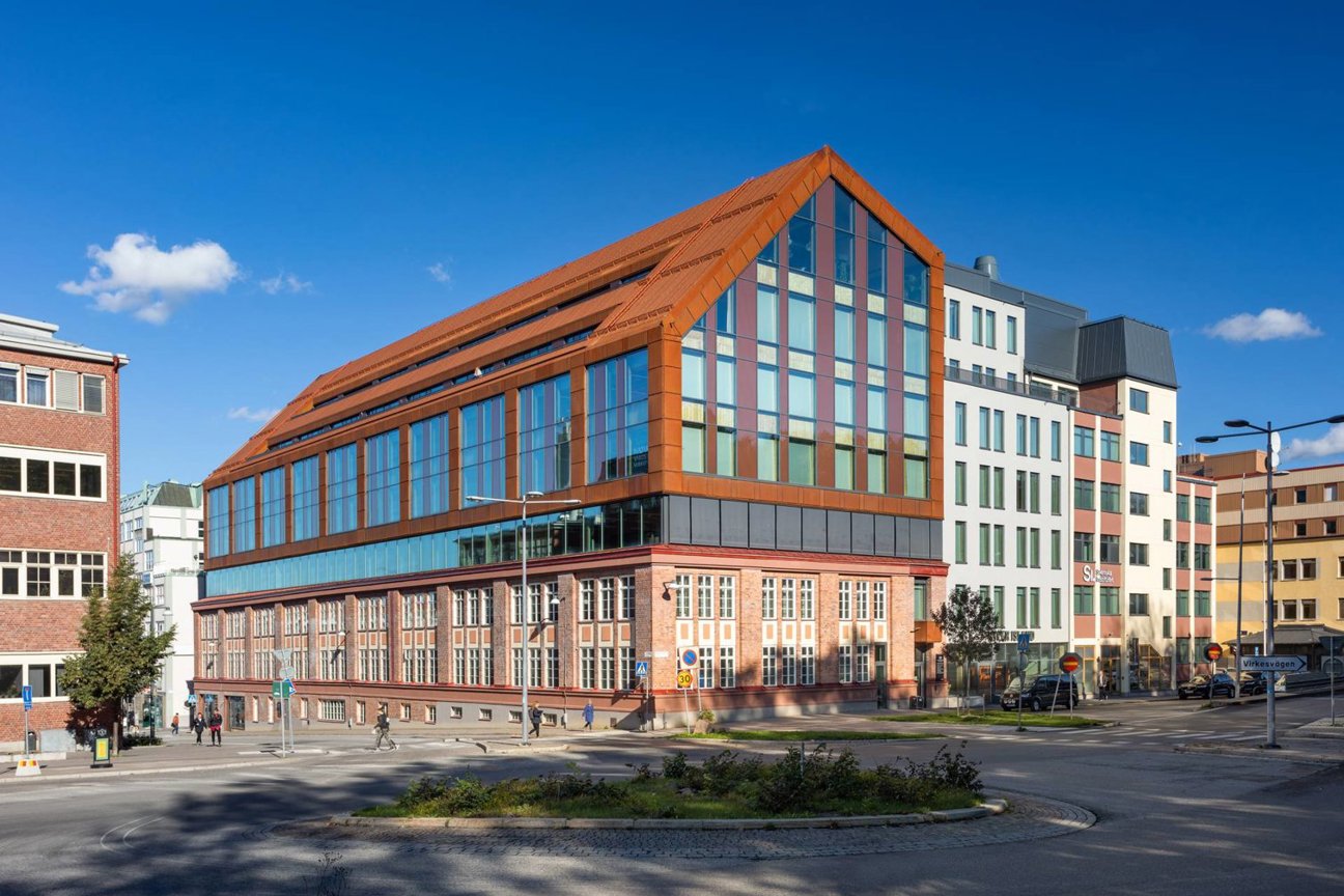 En modern byggnad med rödbrunt tak och stora fönster vid en rundplats under klarblå himmel.