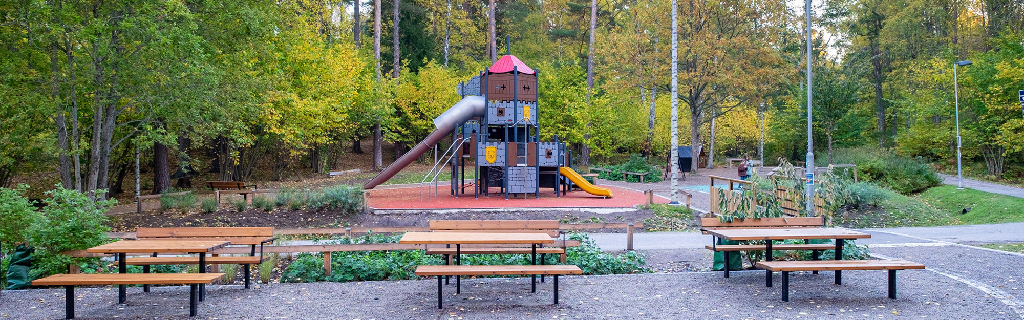 Lekplats med slottformad lekställning, rutschkana och bänkar omgiven av träd.