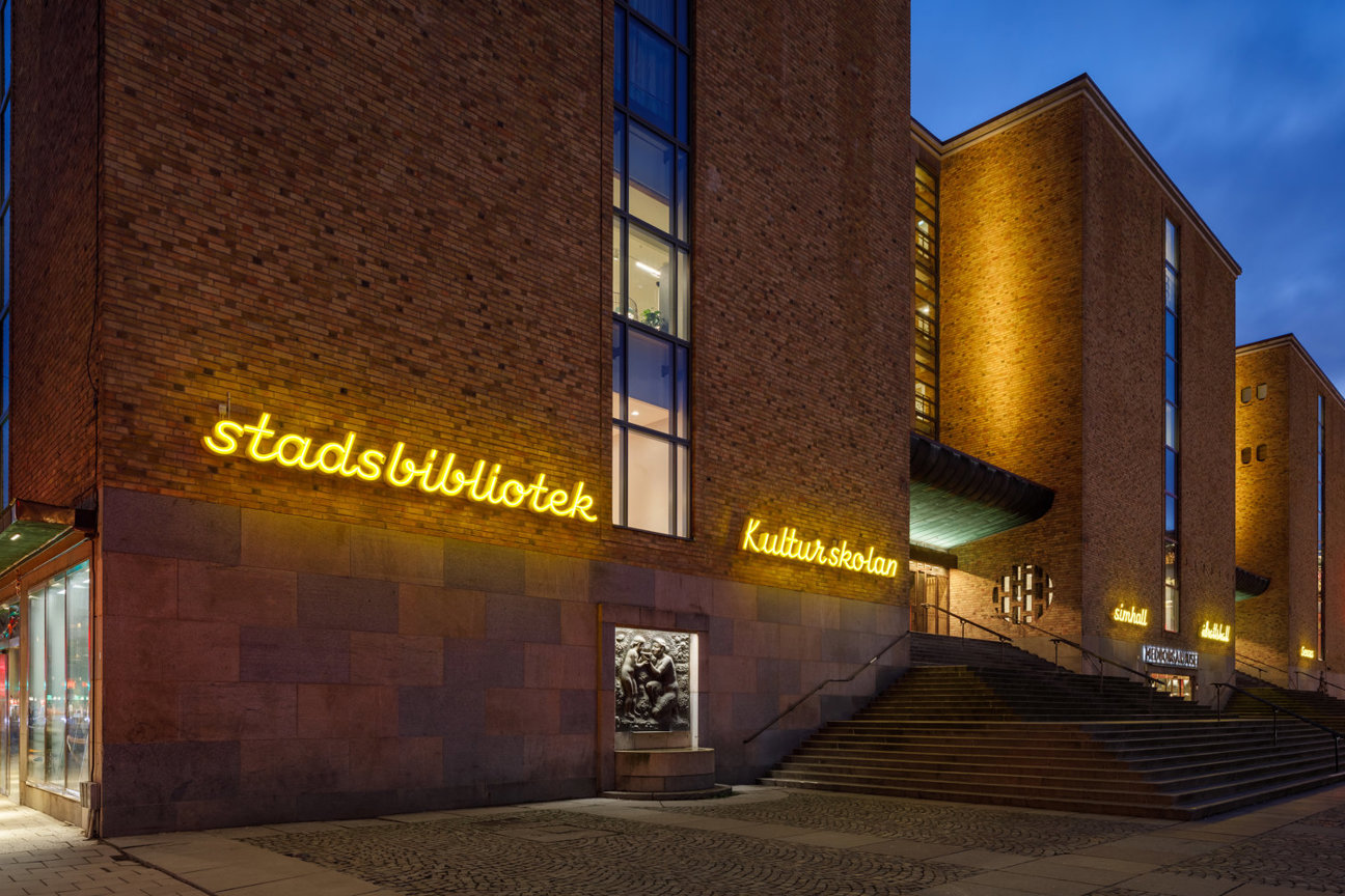 Stadsbibliotekets fasad vid kvällstid med belysta skyltar och trappor.