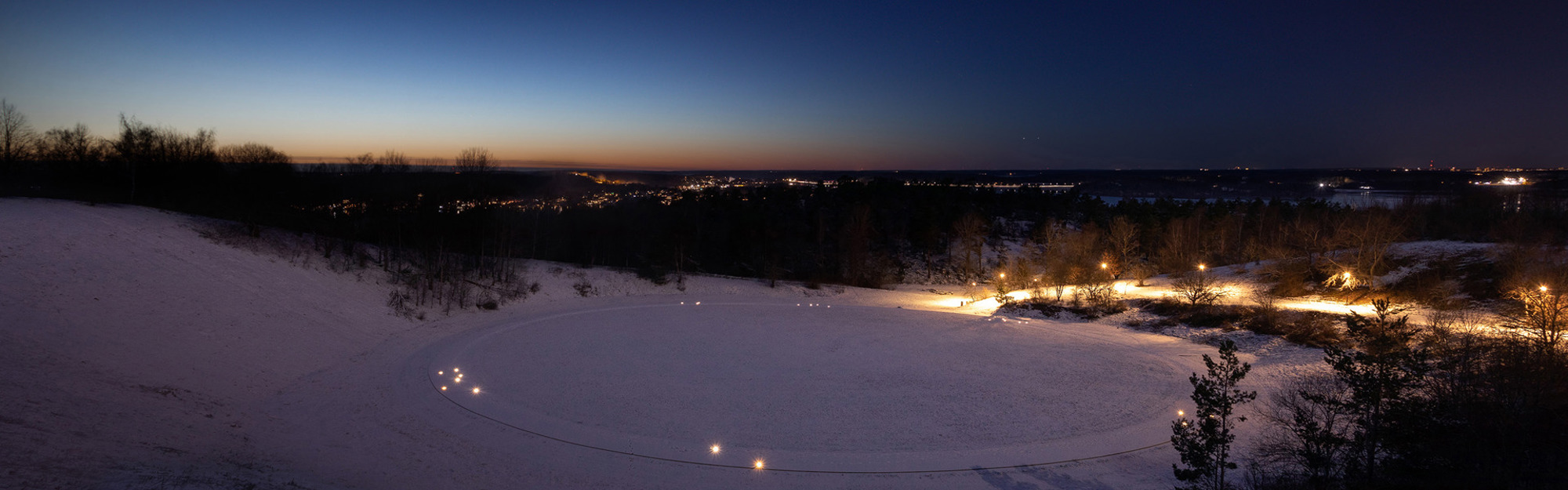 Snöigt landskap vid skymning med cirkulära ljusstråk på snön, träd och belyst stad i bakgrunden.
