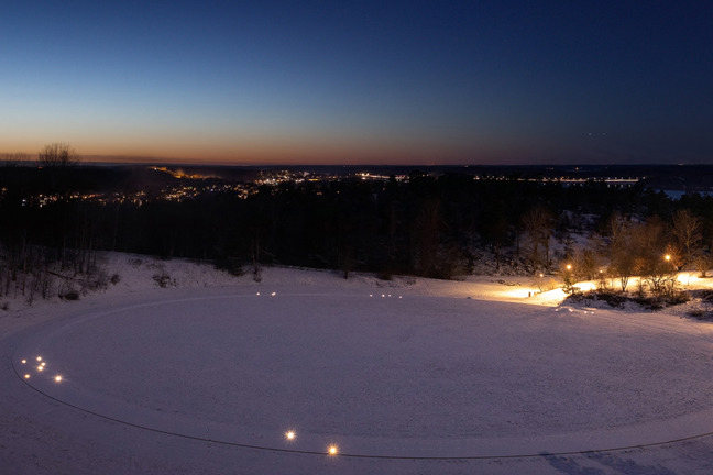 Snöigt landskap vid skymning med cirkulära ljusstråk på snön, träd och belyst stad i bakgrunden.