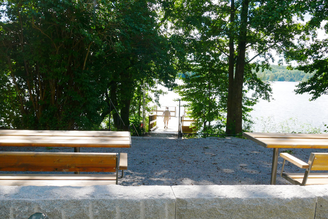 En person går mot en brygga vid en sjö omgiven av träd. Grusväg med träbänkar.