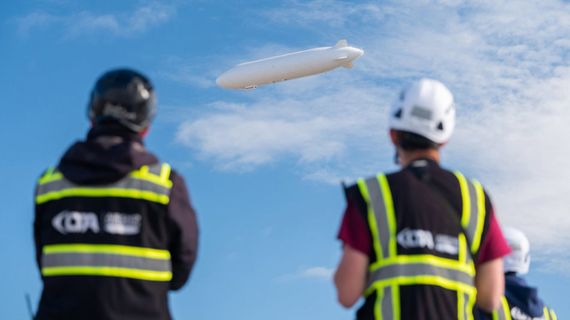 Två personer i reflexvästar och hjälmar tittar på ett luftskepp i blå himmel med moln.