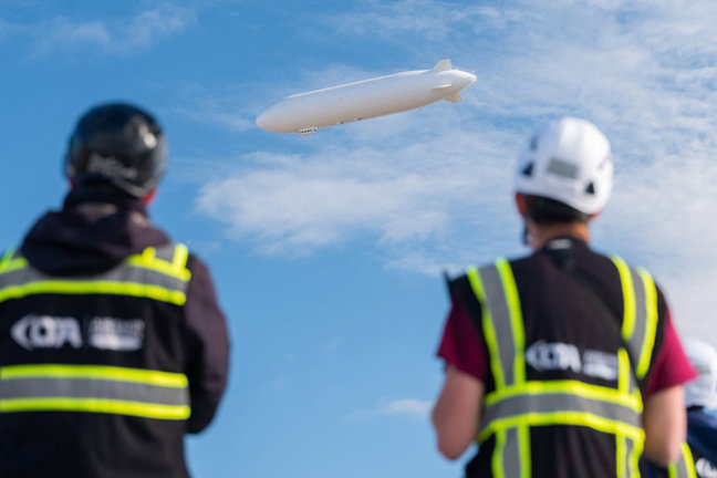 Två personer i reflexvästar och hjälmar tittar på ett luftskepp i blå himmel med moln.