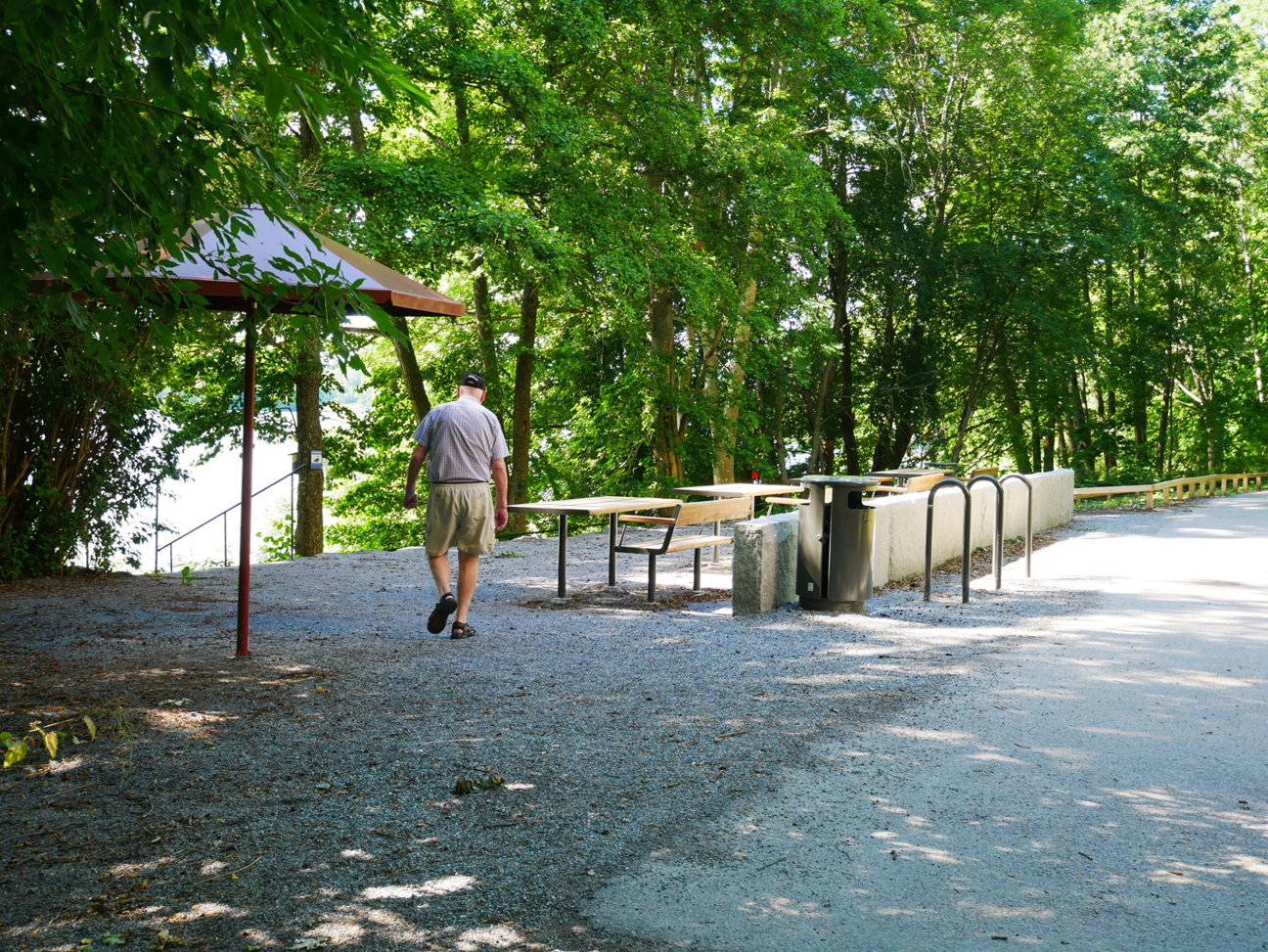 En man går på en grusväg nära bänkar, bord och en paviljong omgiven av träd.