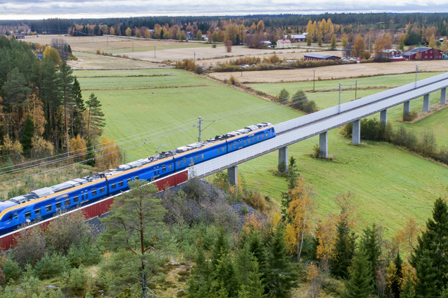 Blå tåg på en viadukt över gröna fält och skogar.