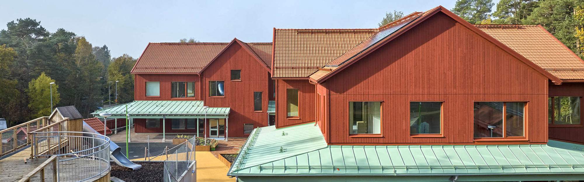 Rött trähus med gröna tak omgivet av träd och lekplats.