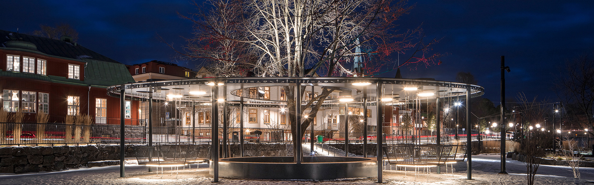 Nattlig torgplats med snöig mark, belysta byggnader, ett blottat träd och en cirkulär ljusstruktur.