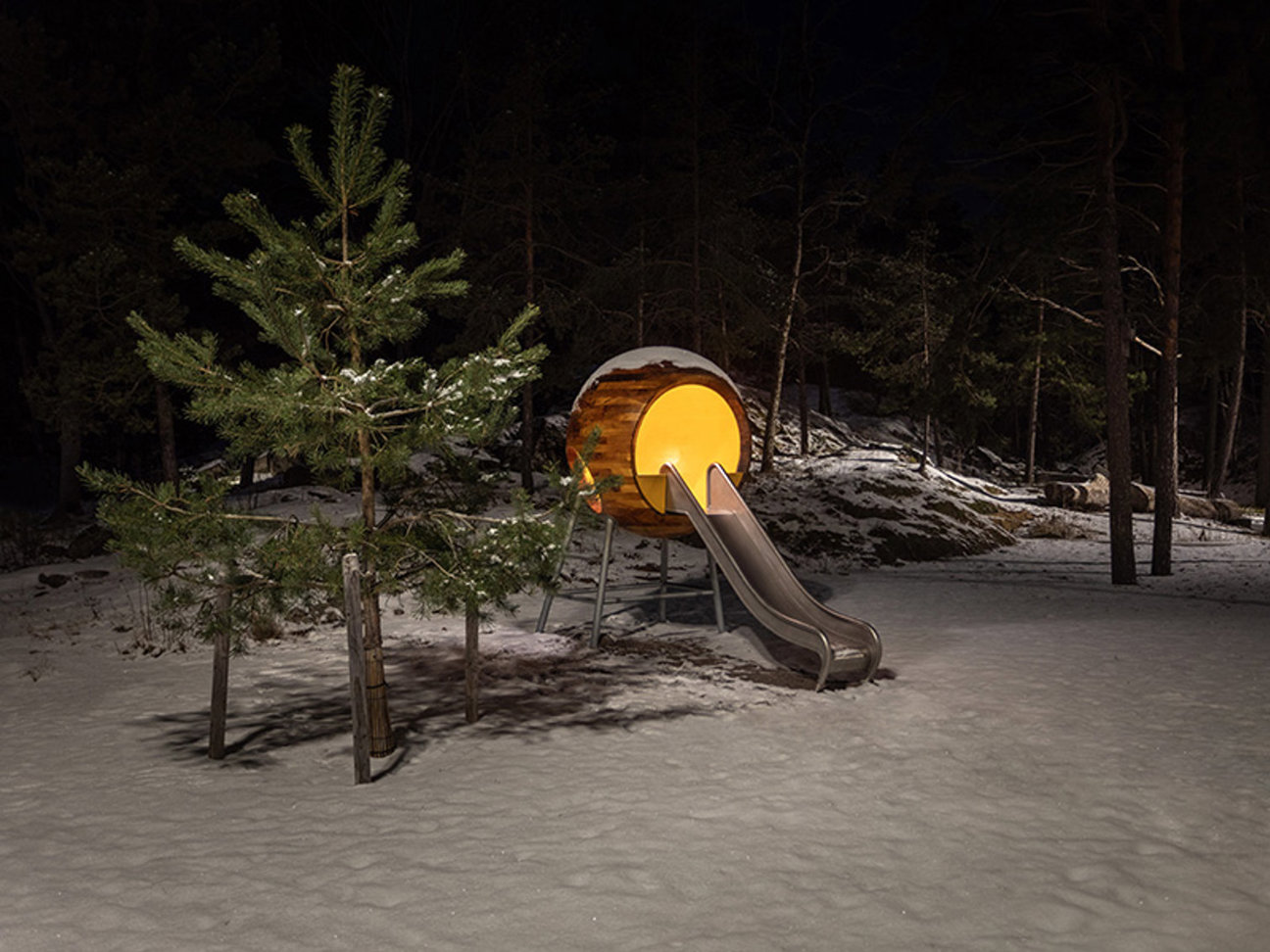 Natt i snöigt skog med en rutschbana ansluten till en upplyst trästruktur och tallar.