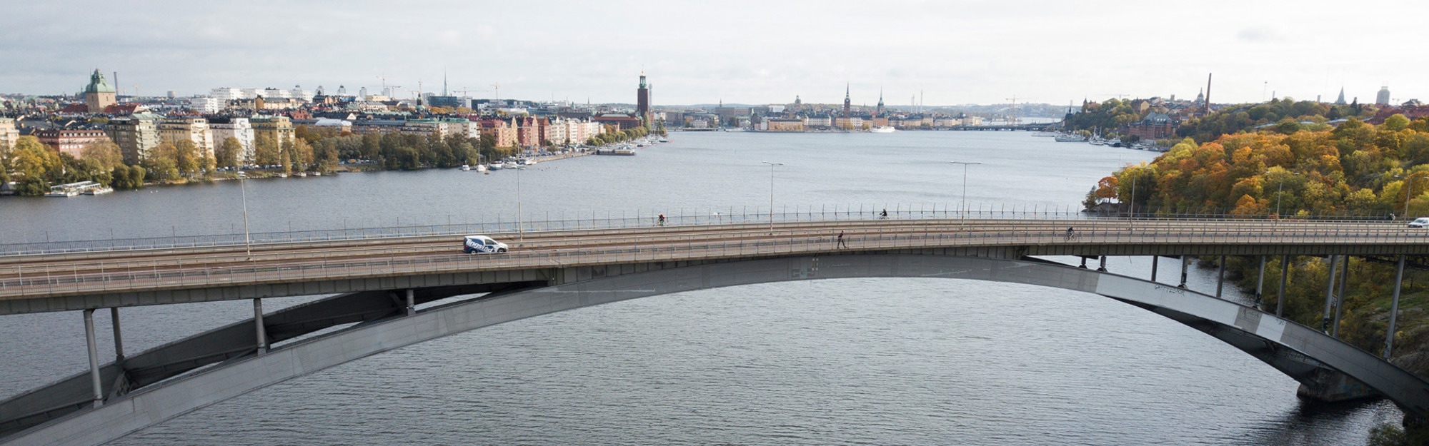 En bro över vatten med stadslandskap i bakgrunden, byggnader och träd med höstfärger.