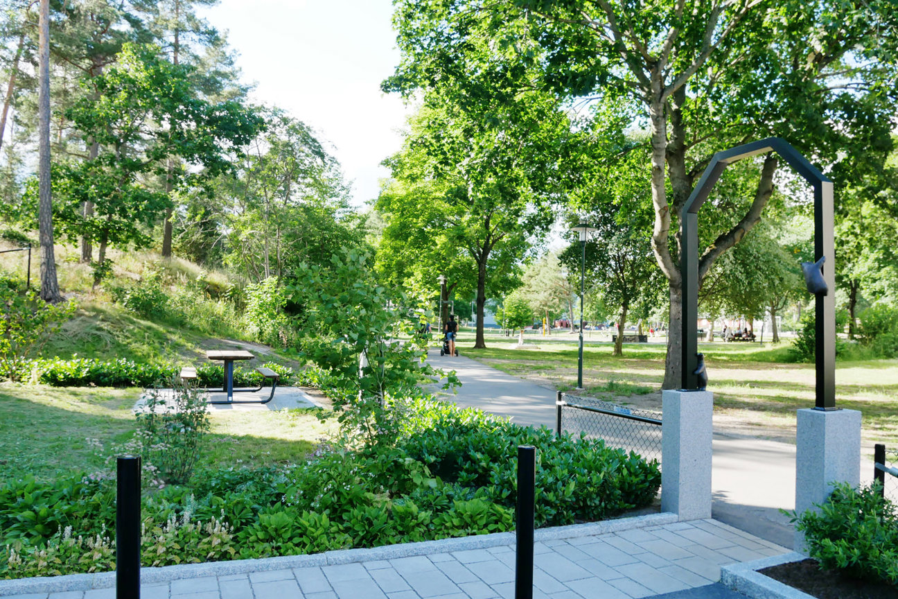 En park med träd, gångväg, bänkar och en person i avståndet.