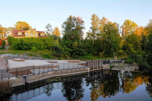 Gul byggnad med röd tak, träd och bro över sjö med spegelbild.