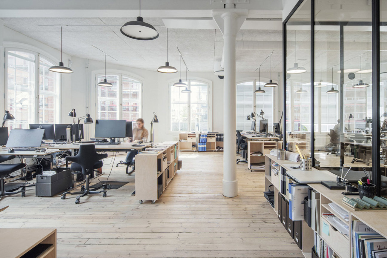 Kontorsutrymme med skrivbord, datorer, stolar och en person vid arbete. Stora fönster och trägolv.