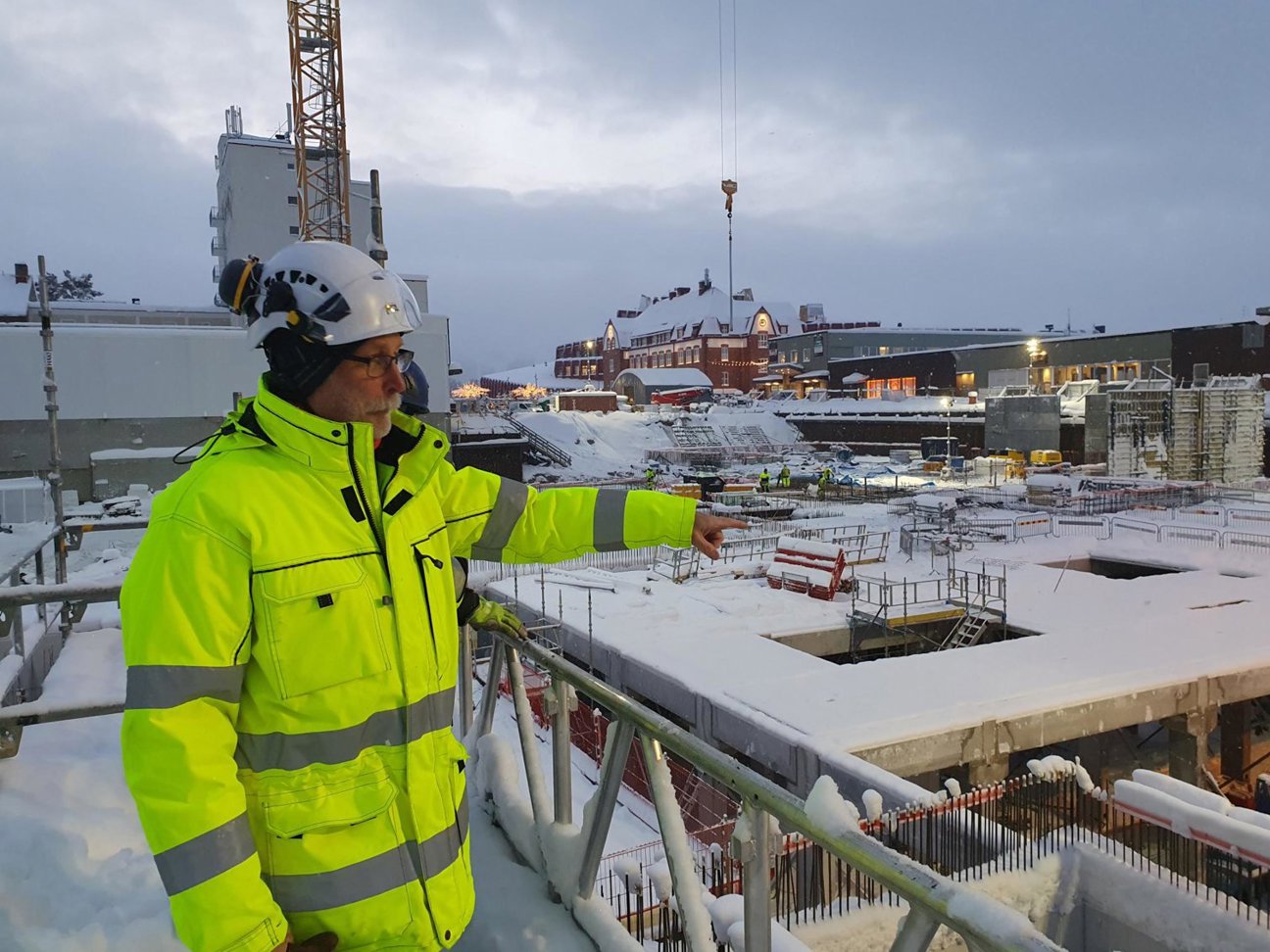 En person i gul reflexväst och vit hjälm pekar på en snöig byggarbetsplats med kranar och byggnader i bakgrunden.