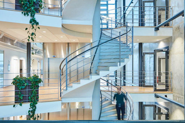 En spiraltrappa i en modern byggnad med en person som går nerför. Växter och glasväggar syns.