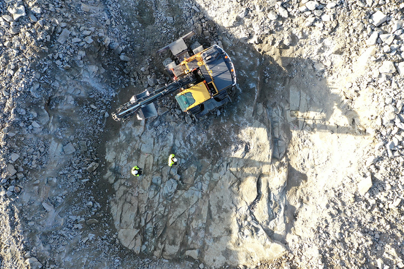 Luftbild av grävmaskin och två personer i säkerhetsvästar på stenig mark.
