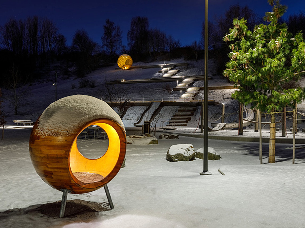 Snöig park med trästrukturer och belysning under mörk himmel.