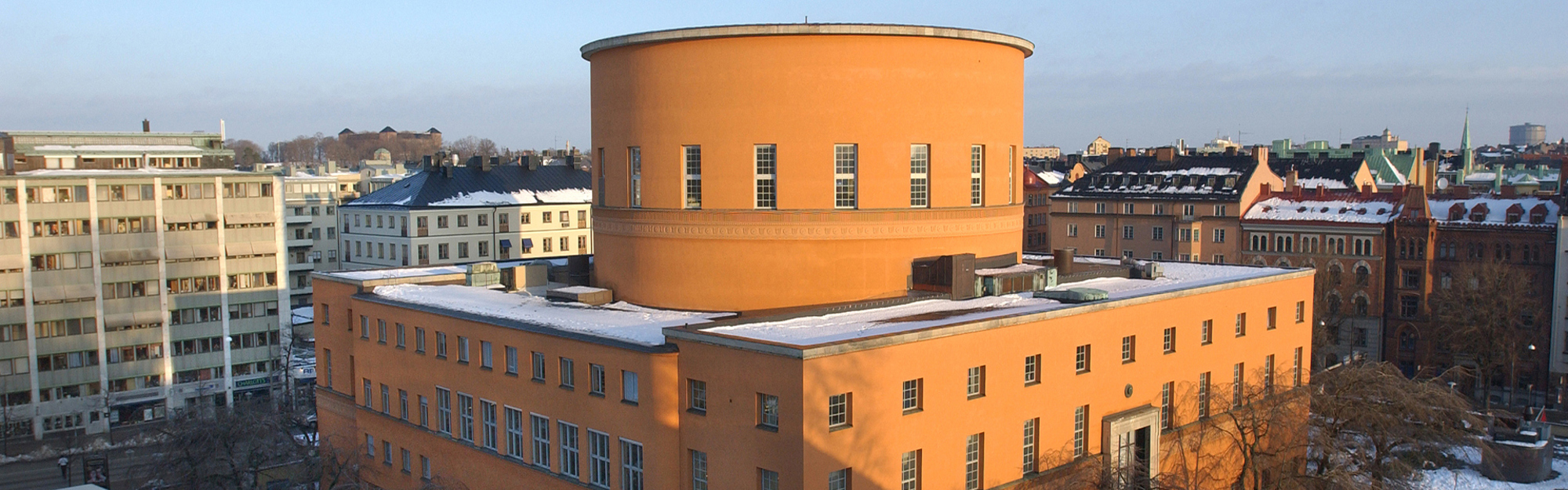 Orange byggnad med cirkulärt torn och snö på tak, omgiven av flera hus i stadslandskap