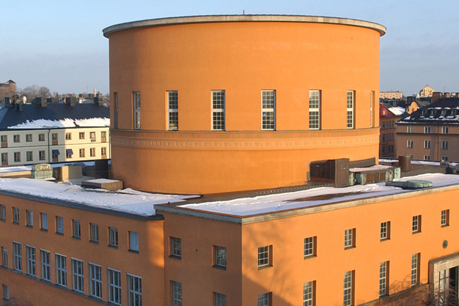 Orange byggnad med cirkulärt torn och snö på tak, omgiven av flera hus i stadslandskap